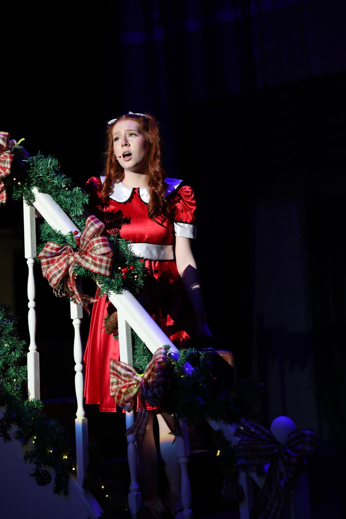 A young actress in Annie's red dress singing on a garlanded staircase under stage light