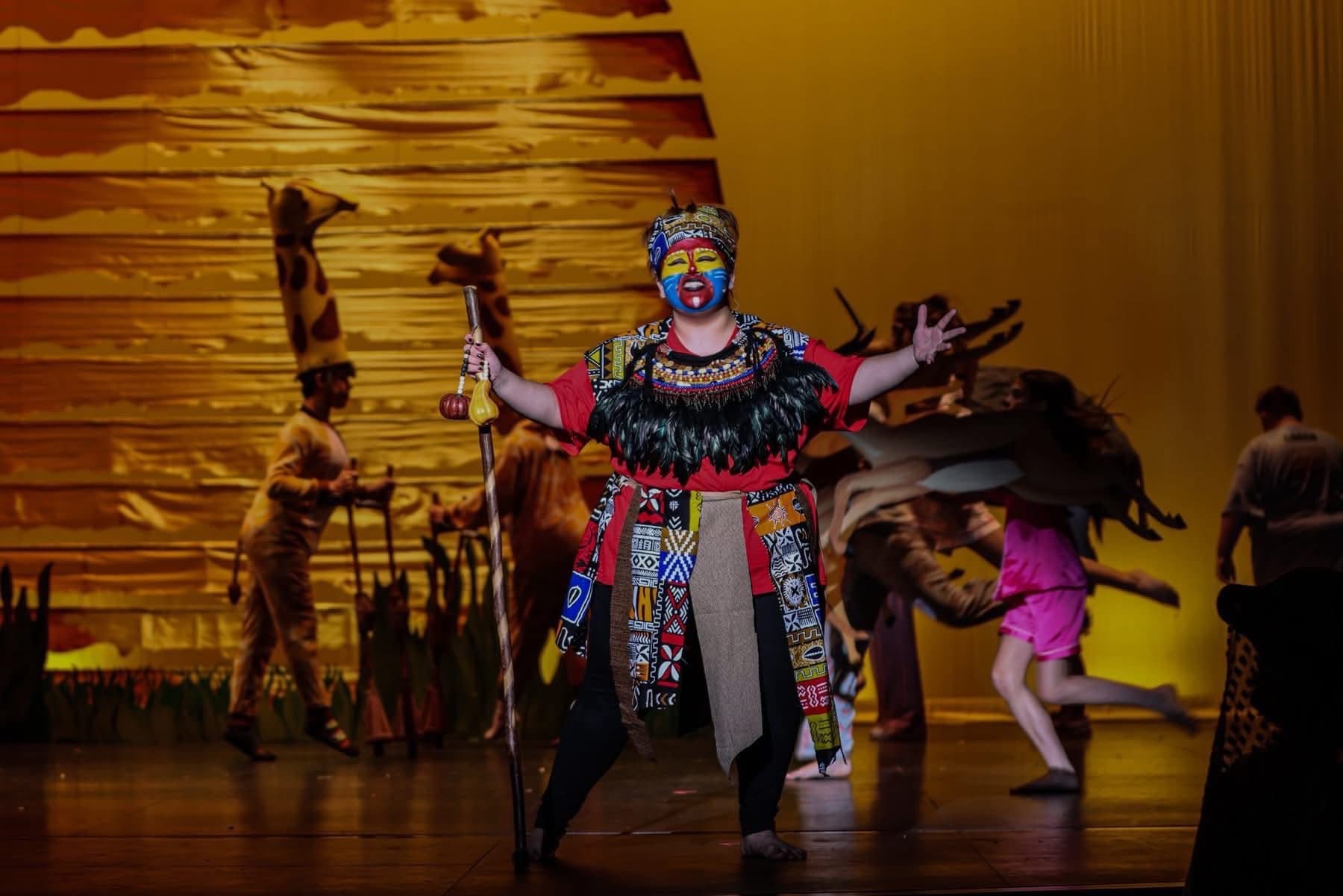 Rafiki in painted mask and feathered costume, arms wide, with the company behind on a lit stage