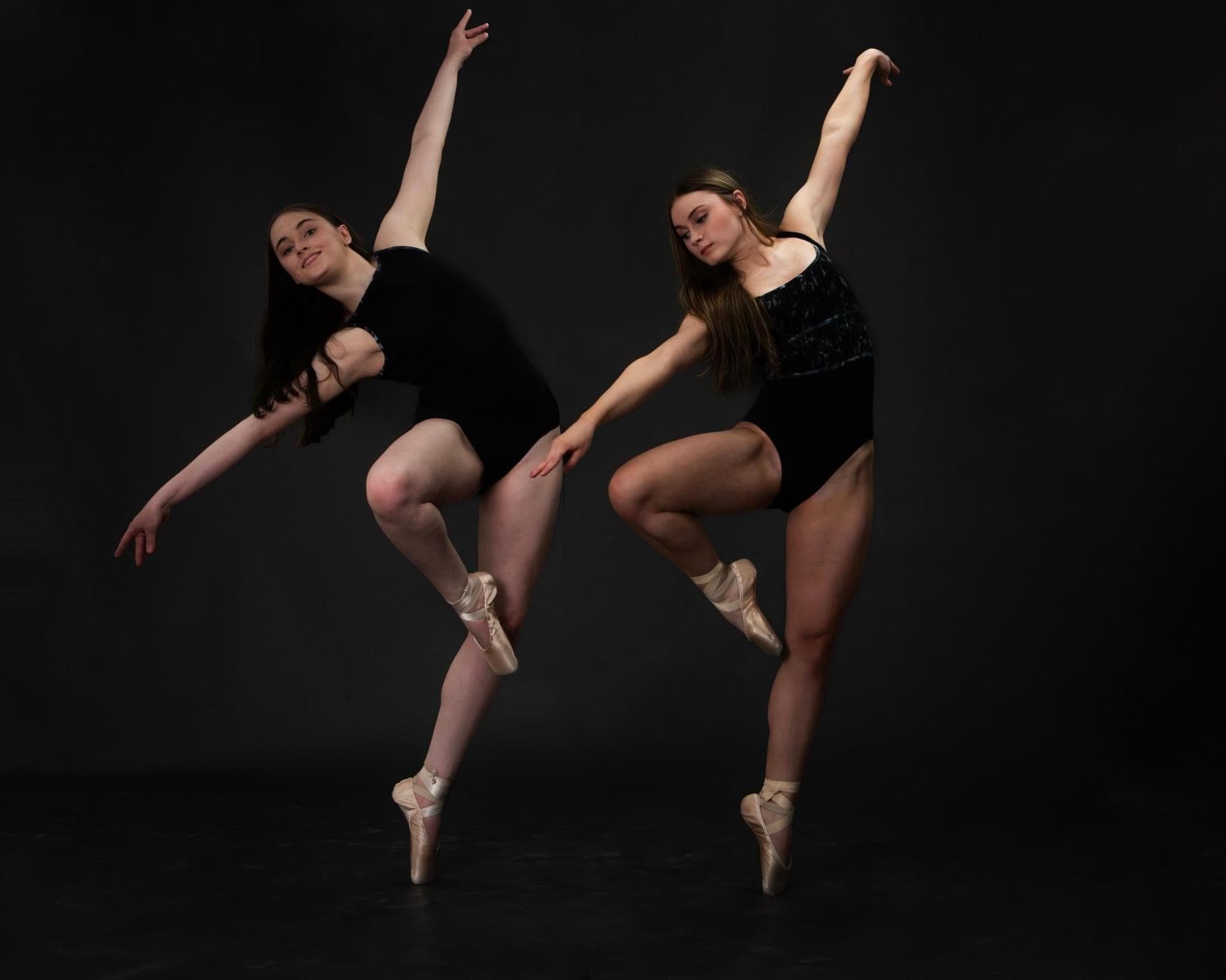 Two dancers en pointe in black leotards posed against a dark backdrop