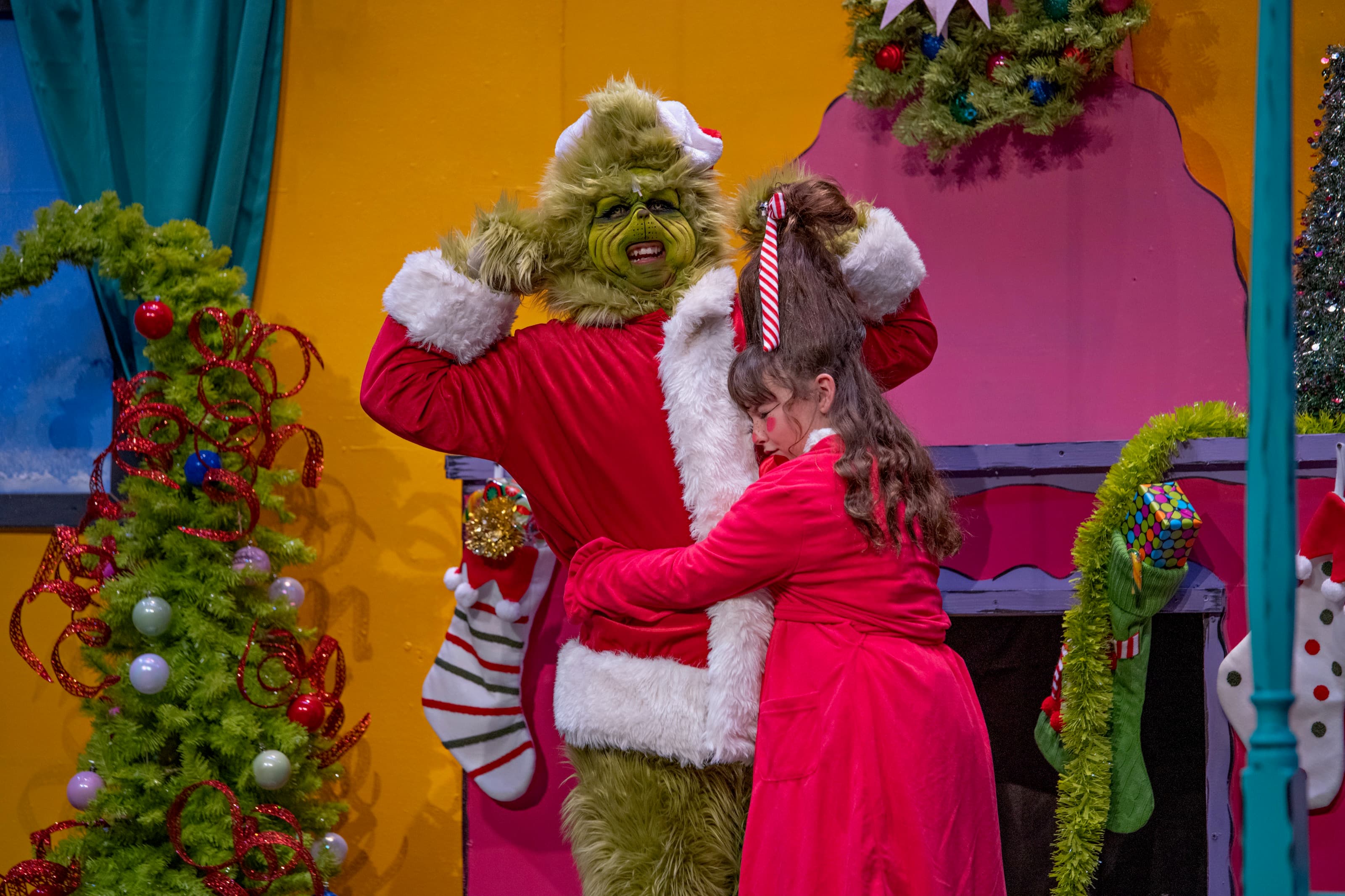 The Grinch in a Santa suit lifting Cindy Lou Who into a hug on a brightly painted Whoville set